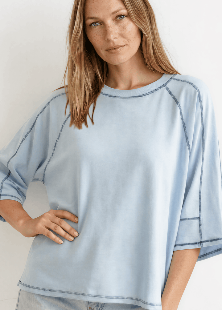  Evelyn Exposed Stitch Tee in Denim Blue. Oversized boxy fit, 3/4 raglan sleeves, contrast topstitching, super soft. Made in USA. Sizes S–XL.
