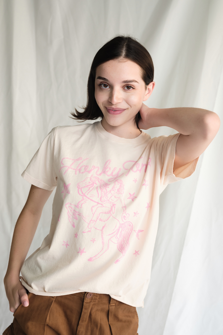 Shop the Honky Tonk Cowgirl Tee — a relaxed-fit crewneck in vanilla white with a pink cowgirl print. 100% cotton, sizes XS–XL.