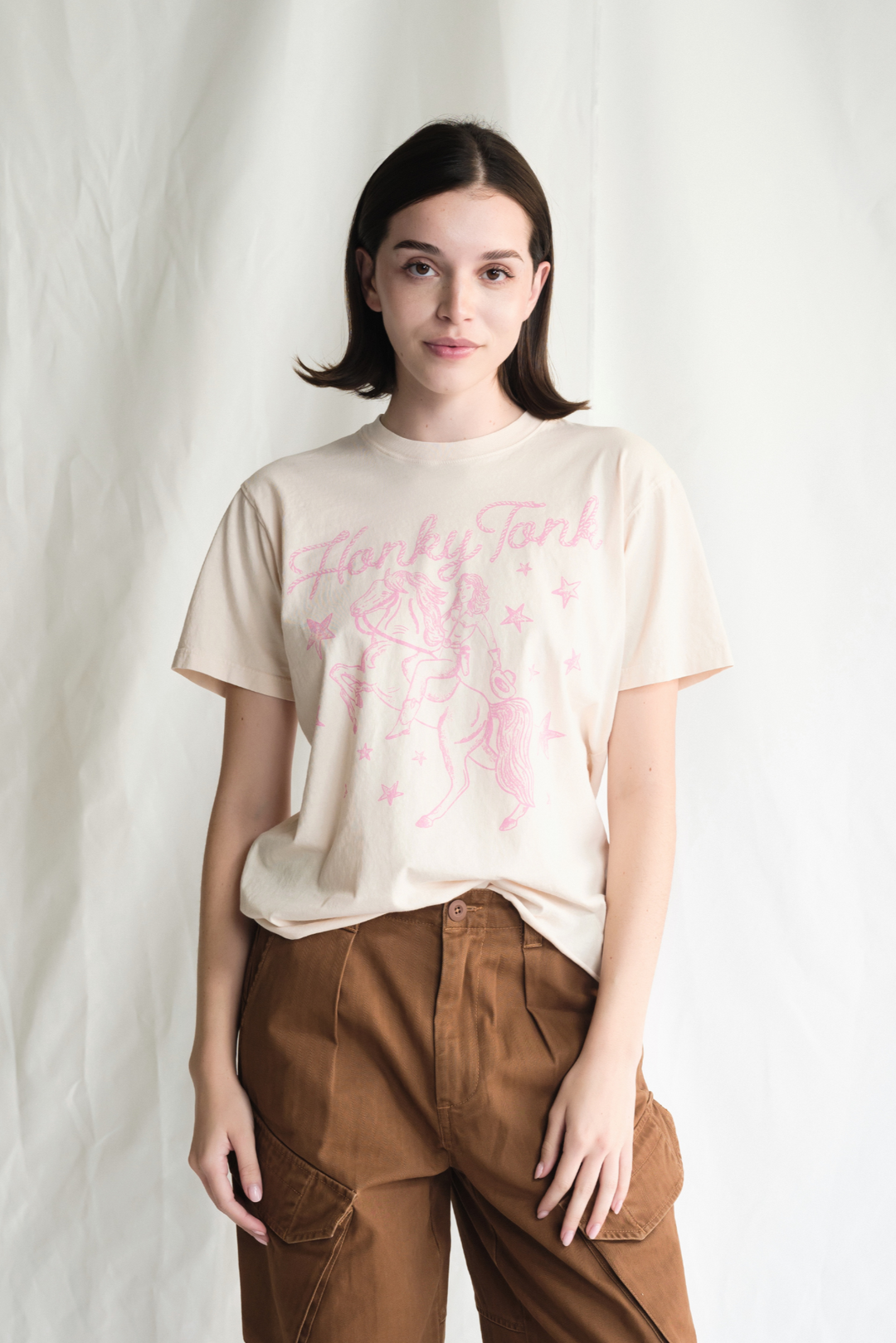 Shop the Honky Tonk Cowgirl Tee — a relaxed-fit crewneck in vanilla white with a pink cowgirl print. 100% cotton, sizes XS–XL.