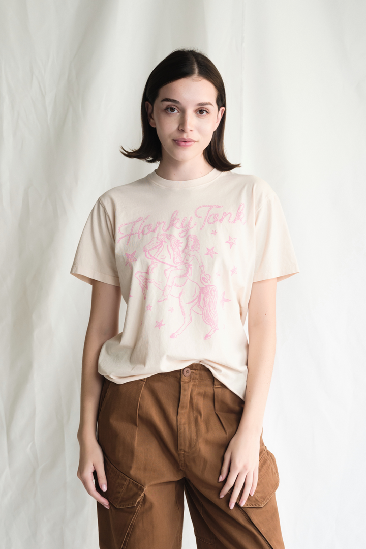 Shop the Honky Tonk Cowgirl Tee — a relaxed-fit crewneck in vanilla white with a pink cowgirl print. 100% cotton, sizes XS–XL.