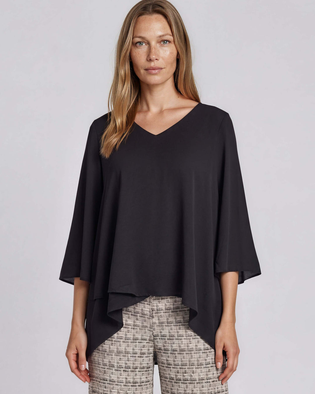 Black dressy V-neck tunic top with mid-length bell sleeves and an overlapping high-low hem that creates a flowy, layered silhouette in lightweight 100% polyester. Made in USA.